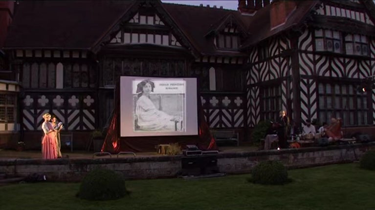A still of a video filmed of the Whose Story? performance at Wightwick in 2007. The still shows a view of the South Terrace of Wightwick Manor in the dark, there is a screen showing an image of one of the Indian Princesses and others are 'on stage' reading from a script and a band is sitting on the right playing traditional South Asian instruments.